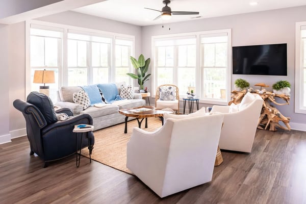 Bright living room with gray sofa, white armchairs, dark leather recliner, wooden coffee table, wall-mounted TV, and large windows overlooking outdoor area.